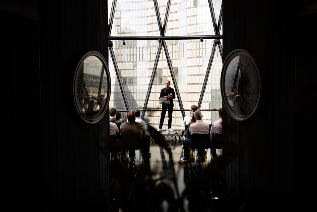Interior of The Gherkin event space set for a conference, with chairs facing a stage and large windows showing London skyline views including Tower Bridge