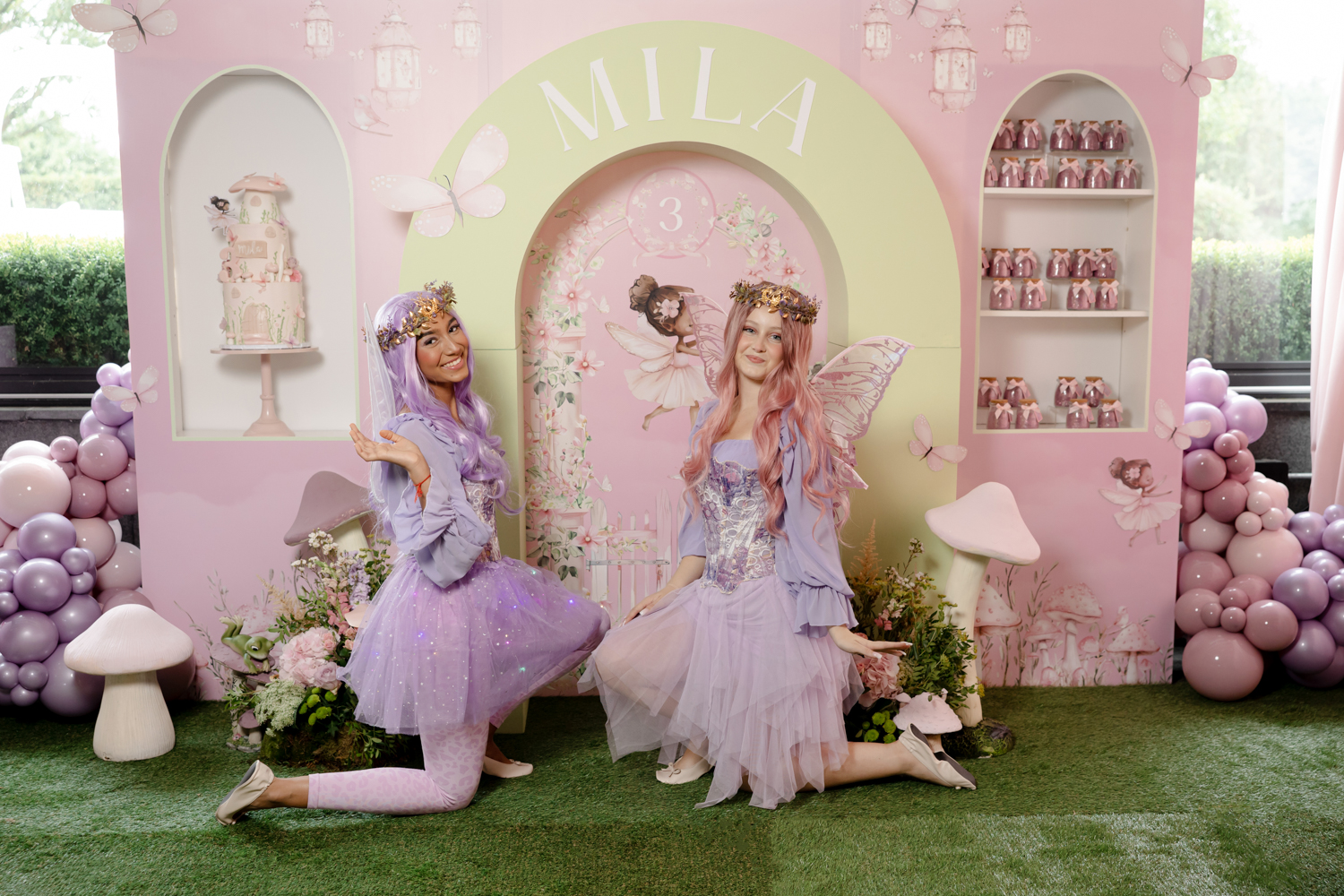 Two entertainers dressed as fairies posing in front of a pink themed birthday backdrop with “Mila 3” sign