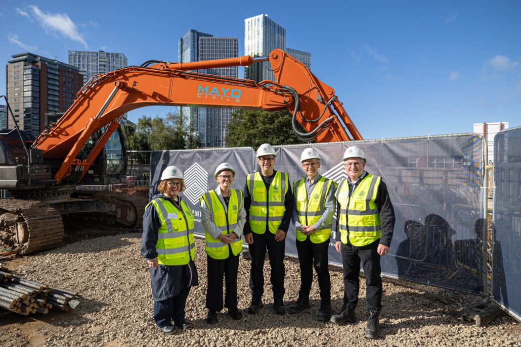 orporate Photography Manchester group photo of stakeholders in high visibility vests and hard hats at Brewery Gardens groundbreaking event with excavator and Manchester skyline.