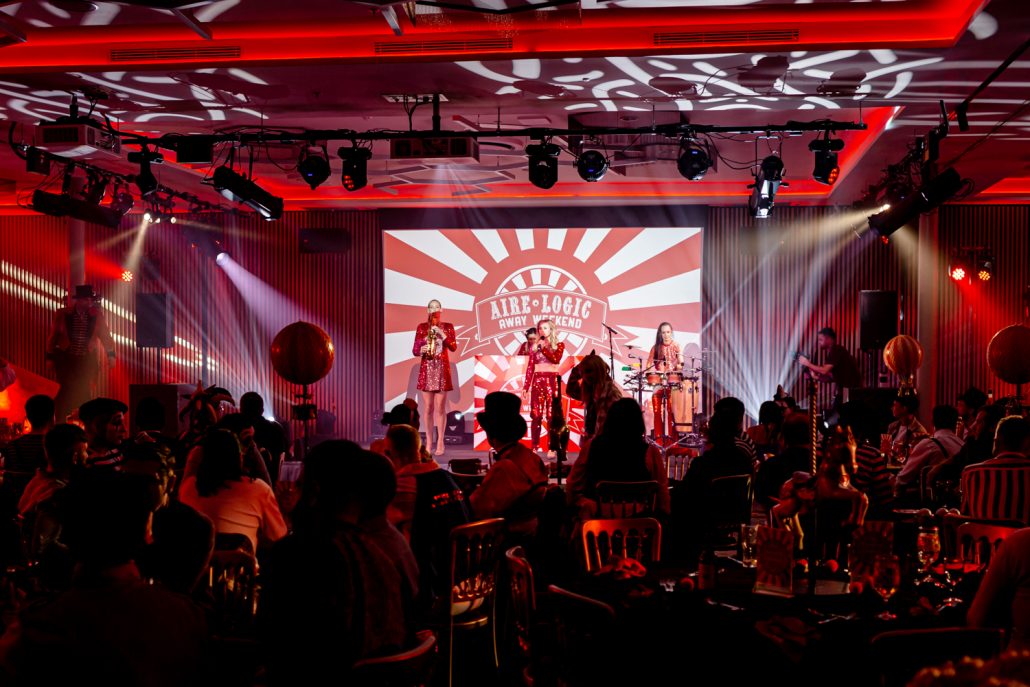 Wide shot of a circus-themed corporate event with live performers on stage, dramatic red lighting, and seated guests enjoying the entertainment at an Aire Logic staff celebration.