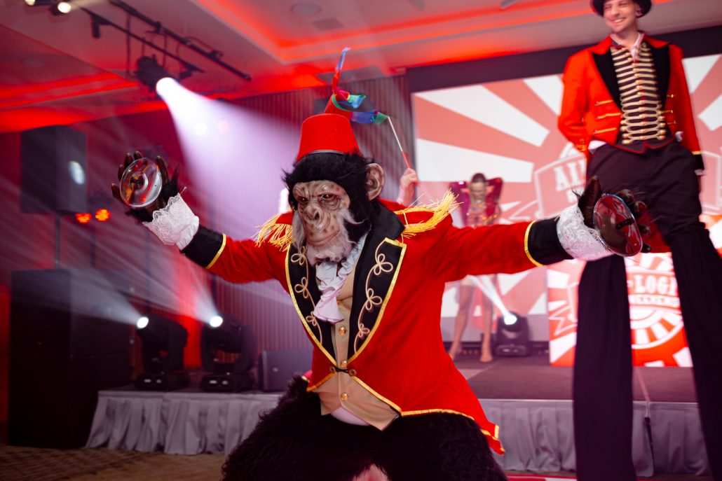 Circus performer dressed as a monkey in a red ringmaster costume playing tambourines in front of the stage during the Aire Logic themed event.